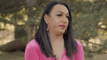 a woman in a pink shirt is standing in front of a tree and looking at the camera .