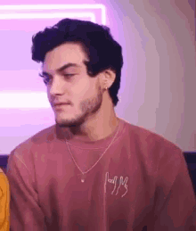 a man with a beard is wearing a pink sweater and a necklace while sitting on a couch .