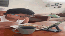 a young boy wearing glasses sits at a table with a plate of food and a mug of coffee