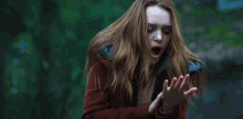 a woman with long red hair is standing in the woods with her hands outstretched and a surprised look on her face .