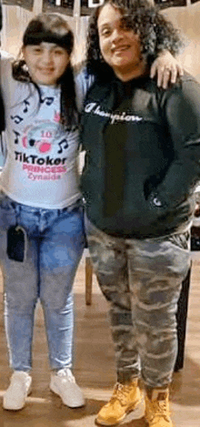 two women are posing for a picture together . one of the women is wearing a tiktoker shirt .