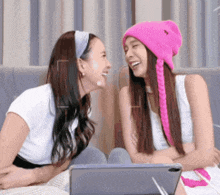 two women are laughing and looking at each other while sitting on a couch . one is wearing a pink hat .