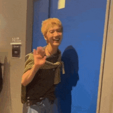 a young man with blonde hair is standing in front of a blue door and waving .