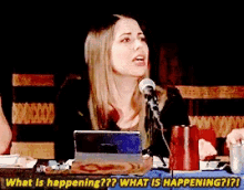 a woman sitting at a table talking into a microphone with the words " what is happening " above her