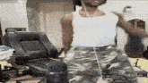 a man in a white tank top and camo pants is dancing in front of a chair .