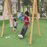 a woman wearing a red adidas shirt is swinging a child on a swing