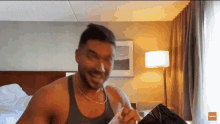 a man in a grey tank top is smiling in a room with a lamp