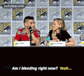 a man and a woman are sitting at a table with microphones in front of a comic con wall .
