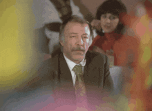 a man in a suit and tie with a woman in a red sweater in the background