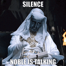 a man in a white robe is sitting in a chair with the words silence noble is talking behind him