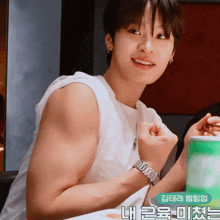 a young man wearing a white tank top and a watch is sitting at a table with korean writing on the bottom