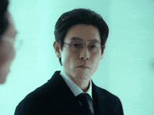 a man wearing glasses and a black suit looks at another man