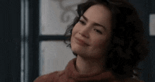 a woman in a red sweater is smiling and looking at the camera in front of a window .