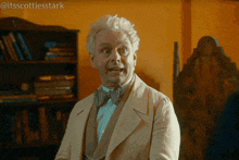 a man with white hair and a bow tie stands in front of a bookshelf with the hashtag @itsscottiesstark