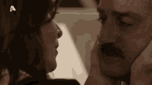 a man with a mustache is being kissed by a woman with the letter a on the bottom right