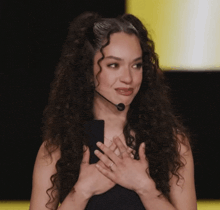 a woman with curly hair wearing a microphone holds her hands to her chest