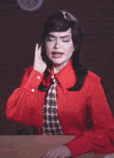 a woman in a red shirt and tie is sitting at a desk with a laptop and the words alert right now below her