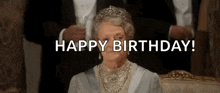 a woman wearing a tiara and pearls is sitting on a couch and says happy birthday .