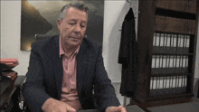 a man in a suit sits at a desk in front of a bookcase