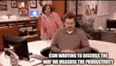 a man is sitting at a desk in front of a computer while a woman walks by .