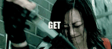 a woman is crying while holding a gun in front of a brick wall with the words `` get '' written on it .