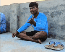 a man in a blue shirt sits on the ground next to a pair of sandals