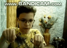 a boy wearing glasses and a yellow shirt is sitting in front of a vase of flowers and making a funny face .