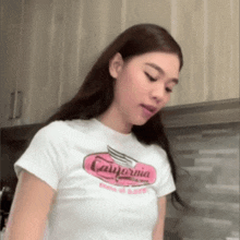 a woman wearing a white california t-shirt stands in a kitchen