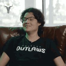 a man wearing a houston outlaws shirt sits on a leather couch
