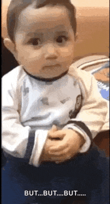 a baby is sitting on a couch and making a funny face while holding his hands together .