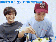 two young men are sitting at a table eating food with chopsticks and smiling .