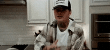 a man wearing a baseball cap and plaid shirt is standing in a kitchen .