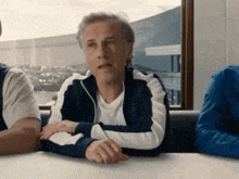 a man in a blue and white tracksuit is sitting at a table with his hands folded .
