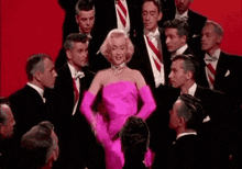 a woman in a pink dress and gloves is standing in front of a group of men in suits and ties .