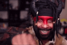 a man with dreadlocks and red paint on his face is wearing a devil costume and pointing at the camera .