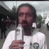 a man with a beard is holding a bottle of water in his hand and saying yes it is .