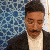 a man with glasses and a mustache is standing in front of a blue and white patterned wall