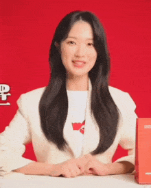 a woman wearing a white jacket and a white shirt with the letter a on it is sitting in front of a red background