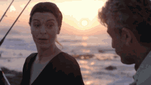 a man and a woman are looking at each other in front of the ocean at sunset