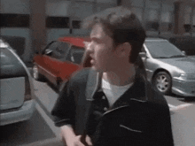 a man in a black shirt is standing in a parking lot next to a red car and a silver car .