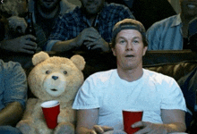 a man sitting on a couch with a teddy bear holding two red cups