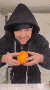 a man in a black hoodie is holding an orange in his hands and smiling .