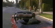 a man is driving a red convertible car down a street with a woman walking behind him .