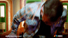 a man in a plaid shirt is standing in front of a sign that says nbc destination x