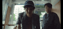 two men are standing next to each other in a hallway . one of the men is wearing a blue hat .