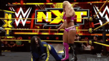 a woman is kneeling down in a wrestling ring with the word nxt on the wall behind her .