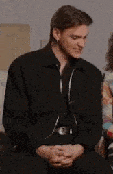 a man in a black jacket is sitting in a chair with his hands folded and smiling .