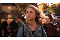 a woman in a denim jacket stands in a crowd of people with her hair blowing in the wind