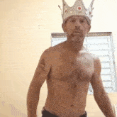 a shirtless man wearing a crown on his head is standing in front of a window .