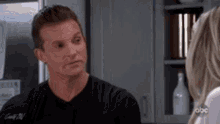 a man is talking to a woman in a kitchen while wearing a black shirt .
